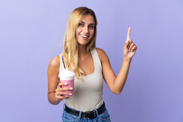 Joven mujer uruguaya rubia con batido de fresa aislado en la pared púrpura apuntando hacia una gran idea