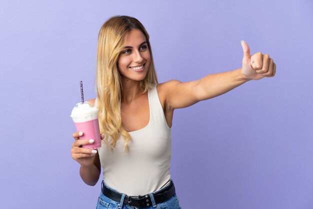 Joven mujer uruguaya rubia con batido de fresa aislado en la pared púrpura dando un gesto de pulgar hacia arriba