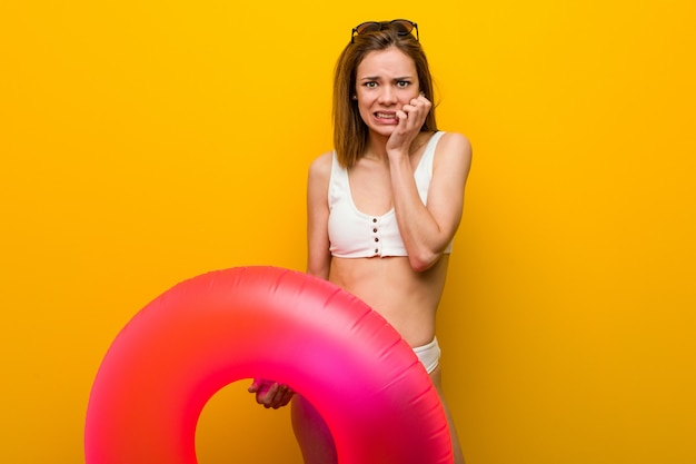 Joven mujer vestida con bikini sosteniendo una rosquilla inflable mordiéndose las uñas, nerviosa y muy ansiosa