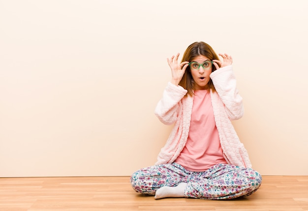 Joven mujer vestida con pijama sentada en casa sintiéndose sorprendida, asombrada y sorprendida, sosteniendo gafas con una mirada asombrada e incrédula