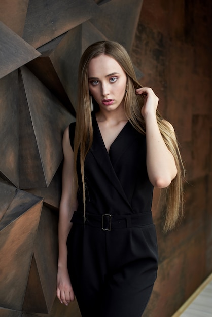 Joven mujer vestida con traje negro, zapatos de tacón posando. Hermosa chica rubia demostrando ropa elegante con estilo contra la pared