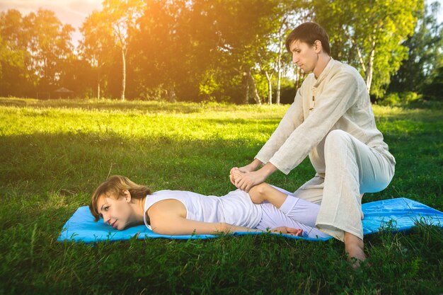 Joven y mujer vestida con una túnica blanca haciendo masaje tailandés con ejercicios de yoga acostado en la colchoneta. Parque de verano soleado con césped verde en el fondo