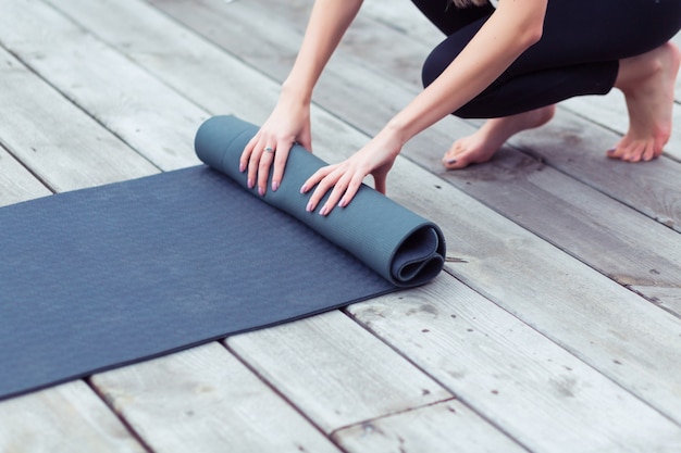 Joven mujer de yoga rodando su estera negra después de una clase de yoga en un piso de madera cerca al aire libre, de cerca