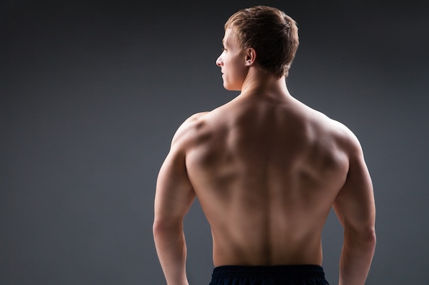 Joven musculoso en estudio sobre fondo oscuro muestra los diferentes movimientos y partes del cuerpo. Vista trasera