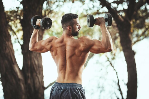 Un joven musculoso en forma con el torso desnudo está haciendo ejercicios con pesas en la naturaleza cerca del río.