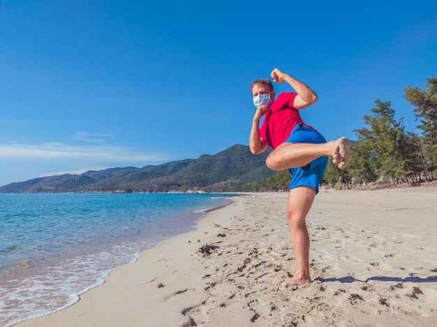 Joven musculoso con máscara médica realiza ejercicios al aire libre en el parque cerca del mar durante la segunda ola de coronavirus Covid 19 cuarentena pandémica Entrenamiento de patada pose brazos oscilantes y piernas deporte vida activa