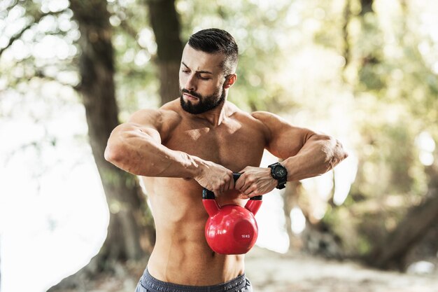 Joven musculoso con el torso desnudo haciendo ejercicios con pesas rusas en la naturaleza.