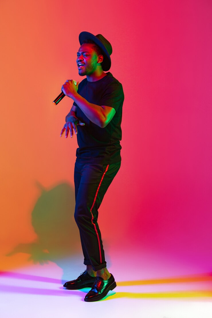 Foto joven músico afroamericano cantando sobre fondo degradado de estudio naranja-púrpura en luz de neón. concepto de música, afición, festival. anfitrión de la fiesta alegre, párese arriba. retrato colorido del artista.