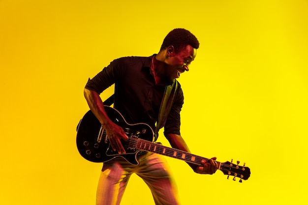 Joven músico afroamericano tocando la guitarra como una estrella de rock en la pared amarilla con luz de neón.