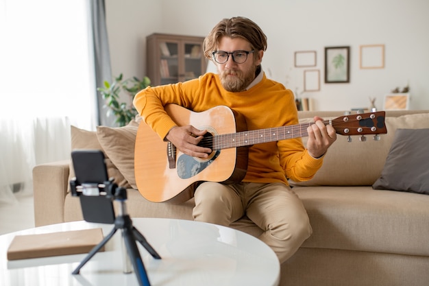 Joven músico barbudo o profesor de música en ropa casual sentado en el sofá en la sala de estar y tocando la guitarra frente a la cámara del teléfono inteligente