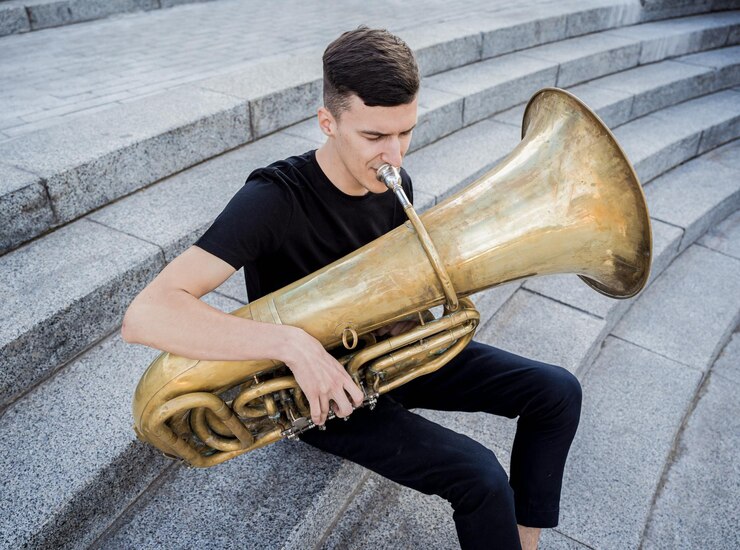 Foto joven músico de la calle tocando la tuba sentado sobre escalones de granito