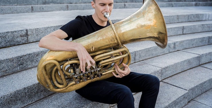 Foto joven músico de la calle tocando la tuba sentado sobre escalones de granito