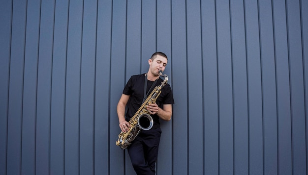 Joven músico callejero tocando el saxofón cerca de la gran pared azul