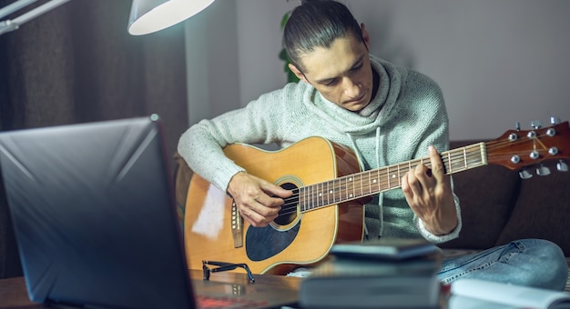 Joven músico está aprendiendo a tocar la guitarra acústica en una lección en línea usando una computadora portátil