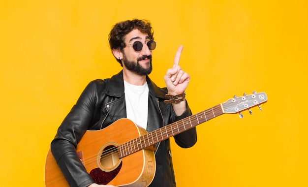 Joven músico sonriendo con orgullo y confianza haciendo la pose número uno triunfante, sintiéndose como un líder con una guitarra