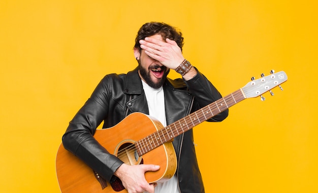 Joven músico sonriendo y sintiéndose feliz, cubriéndose los ojos con ambas manos y esperando una sorpresa increíble con una guitarra