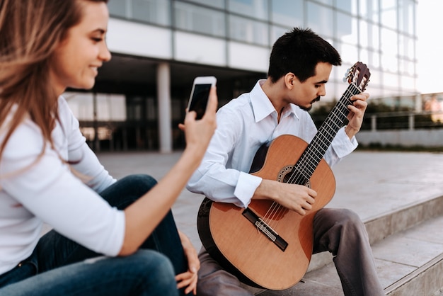 Joven músico tocando la guitarra acústica en la ciudad mientras estaba siendo grabado en un teléfono por una chica.