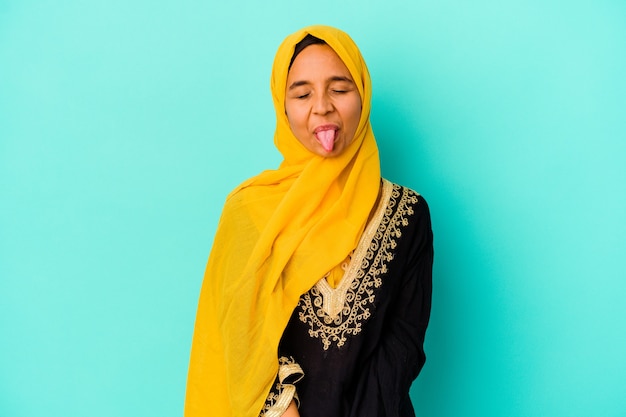 Joven musulmana aislada sobre fondo azul divertido y amigable sacando la lengua.