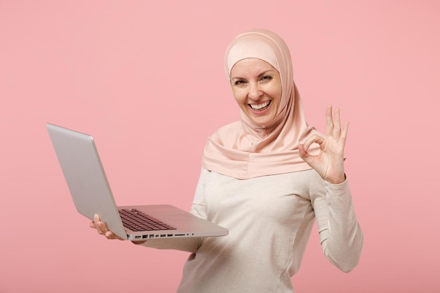 Una joven musulmana árabe alegre con ropa ligera de hijab posando aislada en un fondo rosado. Concepto de estilo de vida religioso de la gente. Simulacros de espacio de copia. Sostenga, trabaje en la computadora portátil, mostrando el gesto OK.