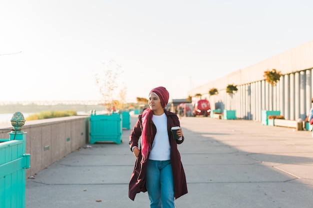 Joven musulmana árabe en hijab caminando en la calle frente al mar y tomando café para llevar copia