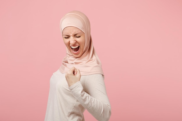 Una joven musulmana árabe muy contenta con ropa ligera hijab posando aislada en un fondo rosado. Concepto de estilo de vida del Islam religioso de la gente. Simulacros de espacio de copia. Gritando, apretando el puño como ganador.