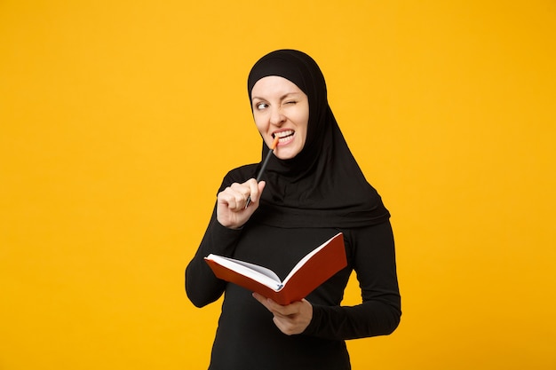 Joven musulmana árabe en ropa negra hijab mantenga cuaderno, escribir, prepararse para el examen aislado en el retrato de la pared amarilla. Concepto de estilo de vida religioso de la gente. .