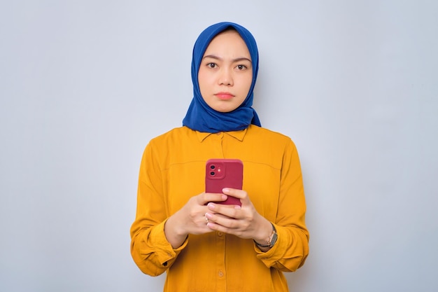 Una joven musulmana asiática ansiosa vestida con una camisa naranja sosteniendo un teléfono móvil y mirando una cámara aislada de fondo blanco