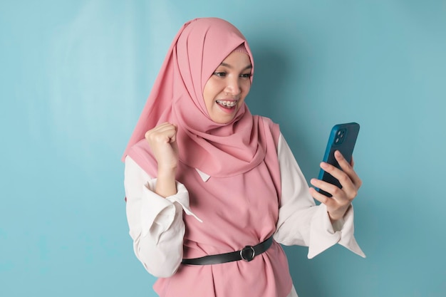 Una joven musulmana asiática con una expresión feliz y exitosa usando hijab rosa y sosteniendo un teléfono inteligente aislado por un fondo azul