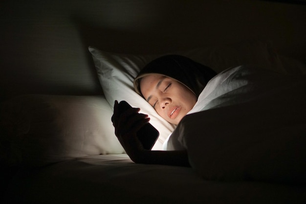 Joven musulmana asiática con un pañuelo en la cabeza mientras está acostada y usa un teléfono móvil por la noche