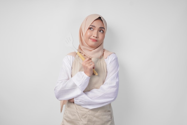 Foto joven musulmana asiática pensativa con hijab y delantal de crema sosteniendo una espátula y utensilios de cocina pensando duro qué menú de comida cocinar