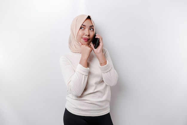 Una joven musulmana asiática pensativa que usa un hiyab y se sostiene la barbilla mientras habla por teléfono aislada por un fondo blanco
