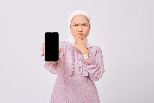 Una joven musulmana asiática seria y hermosa que usa hiyab y un vestido morado que muestra un teléfono inteligente con pantalla en blanco y toca la barbilla aislada en el fondo del estudio blanco