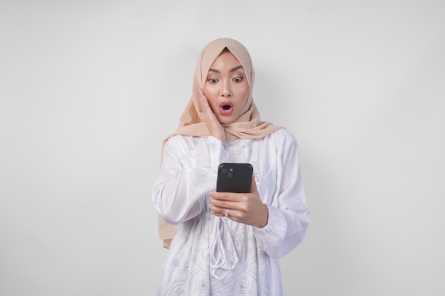 Una joven musulmana asiática con vestido blanco y hijab que muestra una expresión de conmoción en su cara mientras sostiene un teléfono inteligente sorprendida después de leer noticias o chismes aislados por un fondo blanco