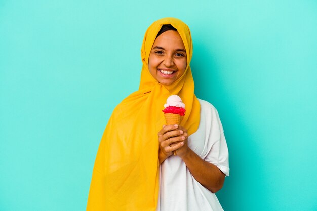 Joven musulmana comiendo un helado aislado sobre fondo azul feliz, sonriente y alegre.