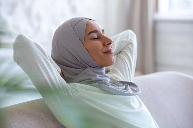 Joven musulmana descansando en casa con los ojos cerrados, las manos detrás de la cabeza, durmiendo la siesta, sentada en el sofá