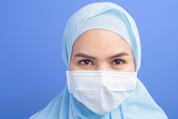 Una joven musulmana con hijab con una mascarilla quirúrgica sobre una pared azul.