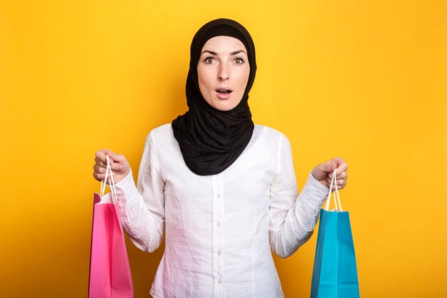 Joven musulmana en hijab tiene bolsas de compras con cara de sorpresa en amarillo