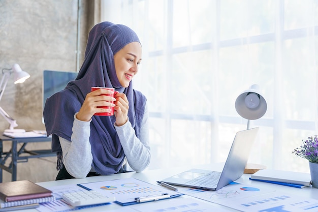 Una joven musulmana que trabaja en una oficina de negocios usando Hijab funciona como un proyecto de PYME de inicio que sostiene una taza de café roja