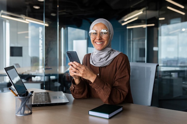 Una joven musulmana sonriente con hijab trabajando en la oficina en una computadora portátil usando un teléfono móvil sonriendo a la cámara