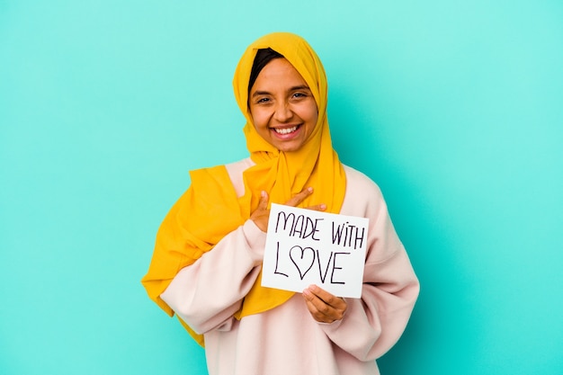 Joven musulmana sosteniendo un cartel hecho con amor aislado en la pared azul se ríe a carcajadas manteniendo la mano en el pecho