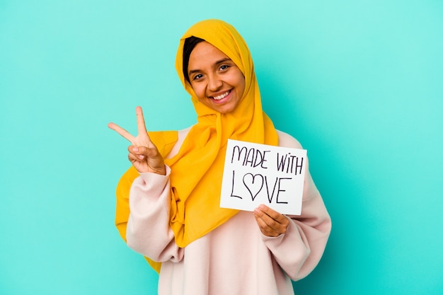 Foto joven musulmana sosteniendo un cartel hecho con amor aislado sobre fondo azul alegre y despreocupado mostrando un símbolo de paz con los dedos.