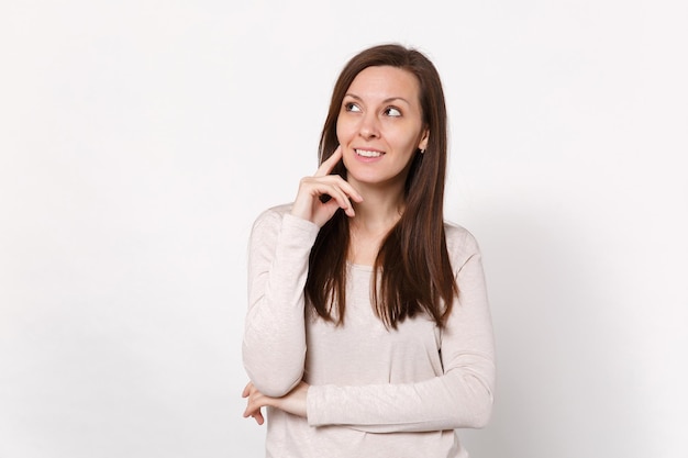 Una joven muy sonriente con ropa ligera mirando a un lado, puso la mano en la barbilla aislada en el fondo de la pared blanca, retrato de estudio. Emociones sinceras de la gente, concepto de estilo de vida. Simulacros de espacio de copia.