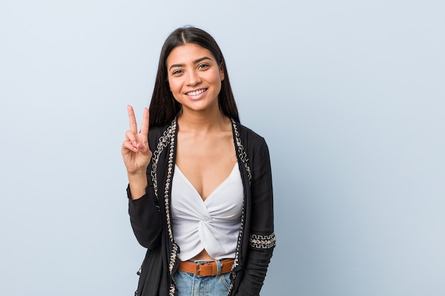 Joven natural y bastante árabe mostrando el signo de la victoria y sonriendo ampliamente.