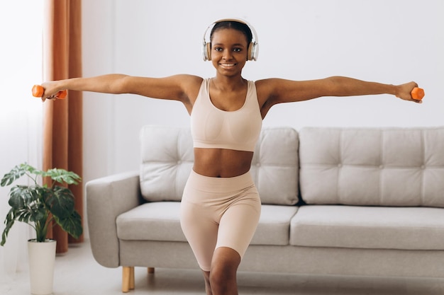 Joven negra con auriculares escucha música y hace ejercicios con pesas en casa en la sala de estar Entrenamiento en casa