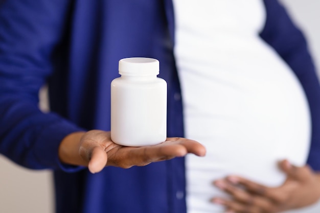 Foto joven negra embarazada con gran barriga muestra un frasco de píldoras de vitaminas en el fondo de la pared blanca