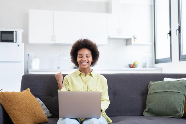 Una joven negra emocionada y alegre que usa una computadora portátil en un sofá en casa recibiendo buenas noticias sintiendo alegría bailando con las manos cantando riéndose haciendo un gesto ganador feliz de ganar un premioxA