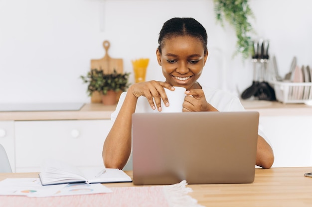 Joven negra sentada en la cocina tomando café y hablando por videollamada en una laptop.