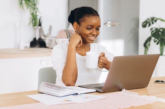 Joven negra sentada en la cocina tomando café y hablando por videollamada en una laptop