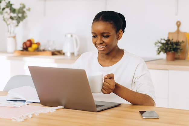 Joven negra sentada en la cocina tomando café y trabajando en una laptop desde casa.