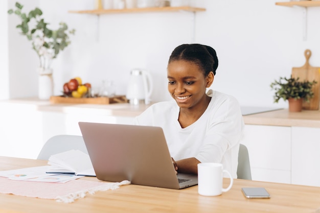 Joven negra sentada en la cocina y trabajando en una laptop desde casa.